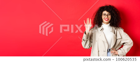 Stylish european woman in glasses and trench coat, waiving hand and smiling, saying hello, greeting someone, standing on red background 100620535