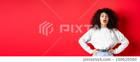Shocked italian woman with curly hair, gasping and staring at camera astonished, open mouth, standing in white sweatshirt on red background 100620560