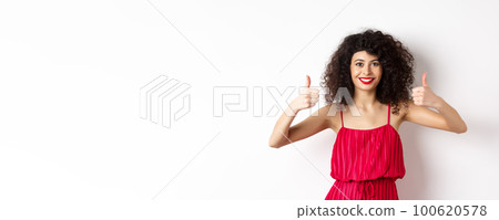 Pretty female model with curly hair and red dress, bright makeup, showing thumbs up in approval, say yes, smiling and nodding in approval, white background Pretty female model with curly hair and red dress, bright makeup, showing thumbs up in approval, say yes, smiling and nodding in approval, white background 100620578