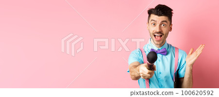Cheerful young man, show host in bow-tie giving you microphone and smiling, entertain people on festive celebration events, performing at holidays, standing on pink background Cheerful young man, show host in bow-tie giving you microphone and smiling, entertain people on festive celebration events, performing at holidays, standing on pink background 100620592