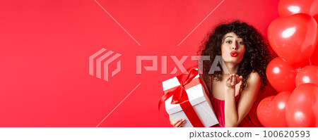 Romance and Valentines day concept. Pretty curly-haired girl in red dress sending her love, blowing air kiss at camera, holding gift from lover, standing near hearts on red background 100620593