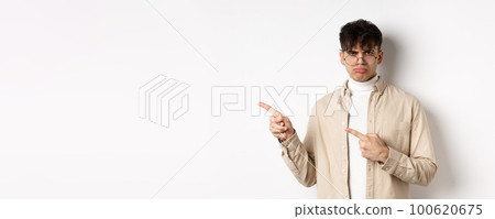 Sad and grumpy boy pointing fingers left, sulking with childish expression, complaining on something bad, standing on white background Sad and grumpy boy pointing fingers left, sulking with childish expression, complaining on something bad, standing on white background 100620675