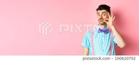 Funny young man touching his french moustache and pucker lips, looking silly at camera, standing in bow-tie and suspenders on pink background 100620702