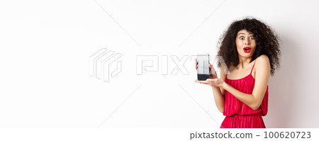 Excited woman with curly hair, makeup and red dress, gasping amazed, showing empty smartphone screen with amazed face, standing against white background 100620723