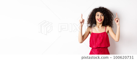 Happy elegant woman in red dress and makeup, smiling amused and pointing fingers up at logo, showing advertisement, standing over white background 100620731