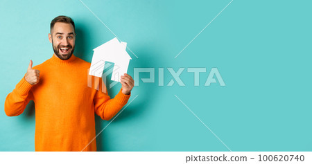 Real estate concept. Satisfied man recommending agency, showing thumb up and house maket, standing over blue background 100620740