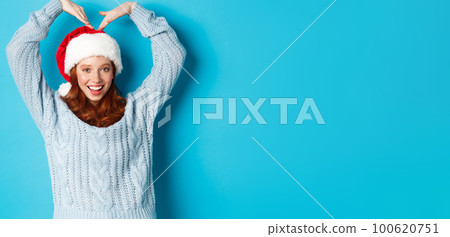Winter holidays and Christmas Eve concept. Cute redhead teen girl in santa hat and sweater, making heart sign and smiling, wishing merry xmas, standing over blue background 100620751