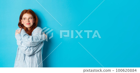 Happy cute redhead girl hugging herself, wearing comfortable and warm sweater, smiling at camera, standing over blue background 100620884