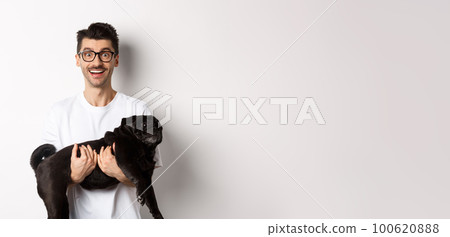 Happy hipster in glasses holding cute black pug and smiling, dog owner staring at camera with amazed smile, standing over white background Happy hipster in glasses holding cute black pug and smiling, dog owner staring at camera with amazed smile, standing over white background 100620888