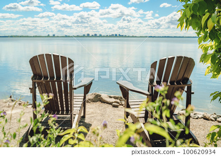 Two Muskoka chairs by the water on home terrace with calm view of lake in Canada. Summer cottage vacation lifestyle Two Muskoka chairs by the water on home terrace with calm view of lake in Canada. Summer cottage vacation lifestyle 100620934