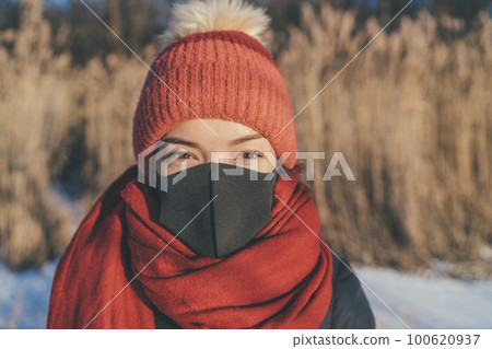 Face mask wearing Asian woman portrait in outdoor winter. Girl in cold weather accessories, hat, scarf for frost bite. 100620937
