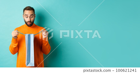 Surprised man open shopping bag and looking amazed, receiving gift on holiday, standing over turquoise background Surprised man open shopping bag and looking amazed, receiving gift on holiday, standing over turquoise background 100621241