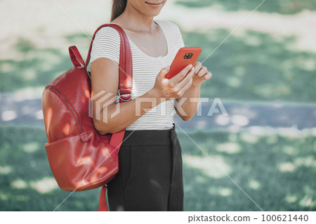 Woman using phone outside walking in park carrying red bag on autumn back to school campus. Online app for vaccine passport. Woman using phone outside walking in park carrying red bag on autumn back to school campus. Online app for vaccine passport. 100621440