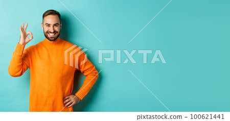Image of confident handsome man showing ok sign, approve and agree, like something good, standing in orange sweater Image of confident handsome man showing ok sign, approve and agree, like something good, standing in orange sweater 100621441