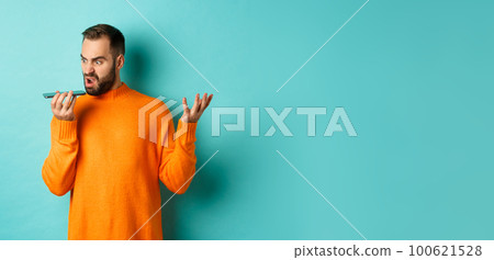 Angry man arguing on speakerphone, record voice message with mad face, standing over light blue background in orange sweater Angry man arguing on speakerphone, record voice message with mad face, standing over light blue background in orange sweater 100621528