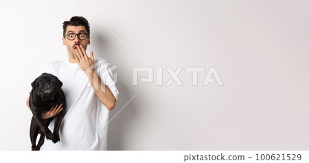 Image of handsome young man holding cute dog and gasping surprised. Pet owner staring at camera shocked, carry black pug in arm, white background Image of handsome young man holding cute dog and gasping surprised. Pet owner staring at camera shocked, carry black pug in arm, white background 100621529