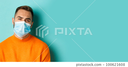 Covid-19, social distancing and quarantine concept. Upset man in face mask complaining, looking displeased and tired, standing against light blue background Covid-19, social distancing and quarantine concept. Upset man in face mask complaining, looking displeased and tired, standing against light blue background 100621608