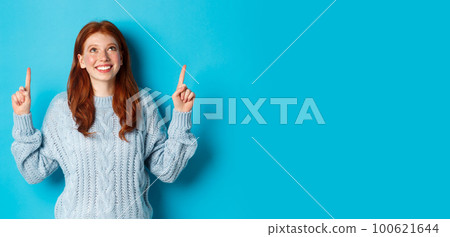 Winter holidays and people concept. Cute teenage redhead girl pointing fingers up, looking at top promo and smiling amused, standing over blue background 100621644