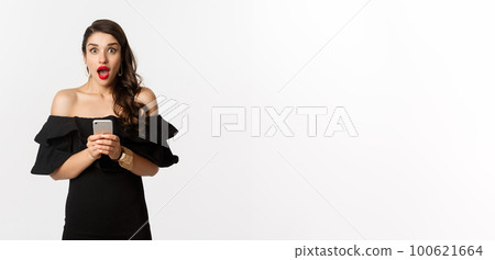 Happy young glamour woman in black dress, holding mobile phone and looking surprised, standing over white background Happy young glamour woman in black dress, holding mobile phone and looking surprised, standing over white background 100621664