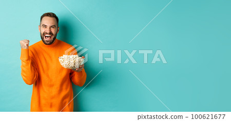 Cheerful handsome man in orange sweater, saying yes, cheering and rejoicing, eating popcorn and watching sports, making fist pump satisfied, standing over blue background Cheerful handsome man in orange sweater, saying yes, cheering and rejoicing, eating popcorn and watching sports, making fist pump satisfied, standing over blue background 100621677