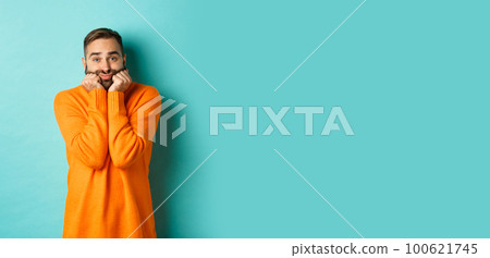 Sad man sighing and looking thoughtful, standing in orange sweater against turquoise background 100621745