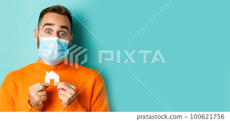 Real estate and coronavirus pandemic concept. Close-up of adult man in medical mask holding small paper house maket and smiling, searching for apartment, light blue background 100621756