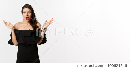 Beauty and fashion concept. Excited beautiful woman looking with amazement at surprise, reacting to good news, standing in black dress with makeup on, white background Beauty and fashion concept. Excited beautiful woman looking with amazement at surprise, reacting to good news, standing in black dress with makeup on, white background 100621846