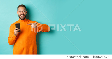 Excited man pointing at mobile phone, showing internet promo, standing over turquoise background, concept of online shopping Excited man pointing at mobile phone, showing internet promo, standing over turquoise background, concept of online shopping 100621872