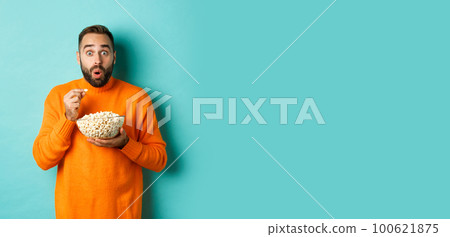 Excited young man watching interesting movie on tv screen, eating popcorn and looking amazed, blue background Excited young man watching interesting movie on tv screen, eating popcorn and looking amazed, blue background 100621875
