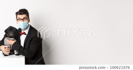 Coronavirus, pets and celebration concept. Happy dog owner in suit and face mask hugging cute black pug in costume, standing over white background Coronavirus, pets and celebration concept. Happy dog owner in suit and face mask hugging cute black pug in costume, standing over white background 100621876
