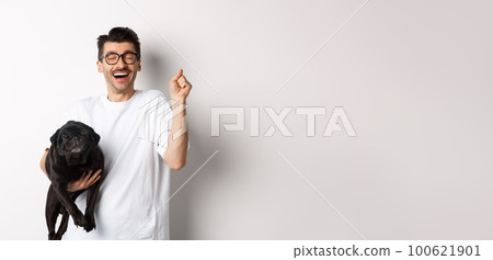 Cheerful handsome man with dog rejoicing, celebrating victory. Happy guy carry cute black pug and looking upbeat, adopting pet, standing over white background 100621901
