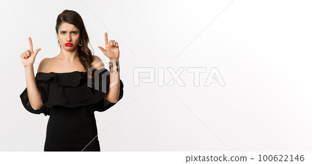 Fashion and beauty. Disappointed woman sulking upset, pointing fingers up and complaining, standing dissatisfied in black dress, white background 100622146