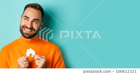 Real estate concept. Happy young man searching for home rent, holding house paper maket and smiling, standing over blue background 100622321