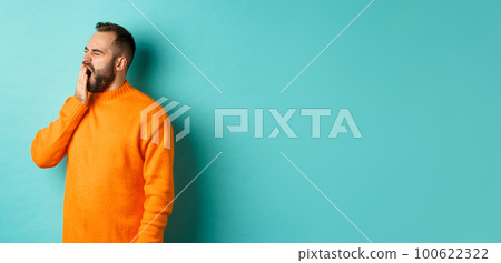Photo of tired bearded man yawning, cover mouth with hand, standing sleepy against light blue background 100622322
