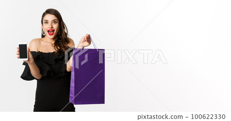Beauty and shopping concept. Beautiful and stylish woman showing smartphone screen and bag, buying online, standing over white background 100622330