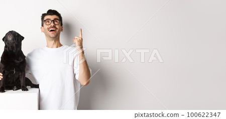 Image of happy young man and dog both looking up at promo, owner pointing finger at top, pug staring at loog, standing over white background 100622347