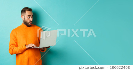 Freelance and technology concept. Image of handsome adult man staring at laptop with amazement, shopping online or working remote, standing over turquoise background Freelance and technology concept. Image of handsome adult man staring at laptop with amazement, shopping online or working remote, standing over turquoise background 100622408