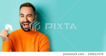 Real estate concept. Happy young man searching for home rent, holding house paper maket and smiling, standing over blue background Real estate concept. Happy young man searching for home rent, holding house paper maket and smiling, standing over blue background 100622476