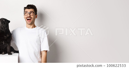 Amazed young man in glasses hugging his dog, pet owner and pug staring at upper left corner promo offer, standing over white background Amazed young man in glasses hugging his dog, pet owner and pug staring at upper left corner promo offer, standing over white background 100622533