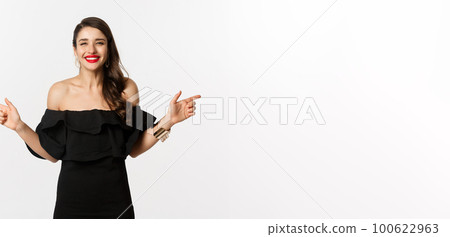 Fashion and beauty. Attractive woman in jewelry, makeup and black dress, smiling and pointing fingers sideways copy space offer, white background Fashion and beauty. Attractive woman in jewelry, makeup and black dress, smiling and pointing fingers sideways copy space offer, white background 100622963