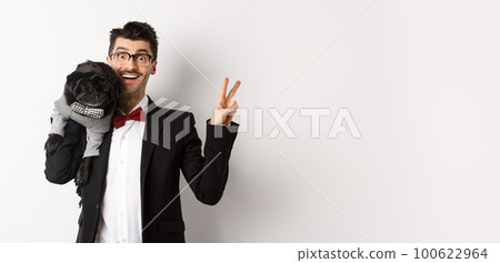 Cheerful young man in suit and glasses taking photo with cute black pug dog on his shoulder, smiling happy and showing peace sign, posing over white background Cheerful young man in suit and glasses taking photo with cute black pug dog on his shoulder, smiling happy and showing peace sign, posing over white background 100622964