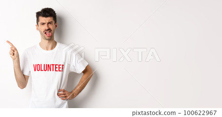 Disappointed volunteer showing bad promo offer, sticking tongue and grimacing disgusted, pointing finger left, wearing organization t-shirt, white background Disappointed volunteer showing bad promo offer, sticking tongue and grimacing disgusted, pointing finger left, wearing organization t-shirt, white background 100622967