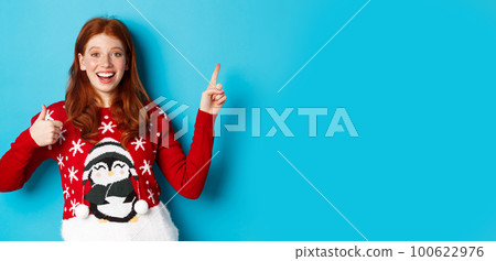 Merry Christmas. Cheerful redhead girl in xmas sweater, pointing finger at upper right corner, showing new year promo and thumbs-up in approval, praise product 100622976