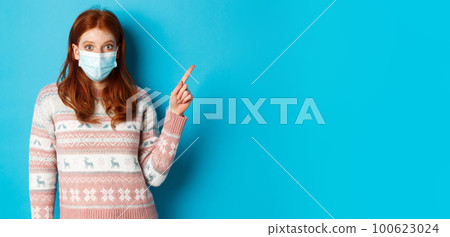 Winter, covid-19 and quarantine concept. Intrigued redhead girl in medical mask picking product, looking and pointing at upper right corner promo, blue background 100623024