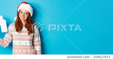 Holiday promos and real estate concept. Cheerful redhead woman in santa hat holding paper house in hand and smiling, standing in sweater against blue background 100623033