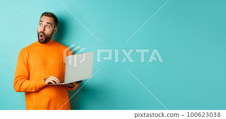 Handsome man doing online shopping, looking up thoughtful while using laptop, standing over light blue background Handsome man doing online shopping, looking up thoughtful while using laptop, standing over light blue background 100623038