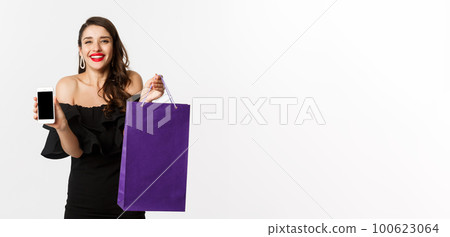 Beauty and shopping concept. Beautiful and stylish woman showing mobile phone screen and bag, buying online, standing over white background Beauty and shopping concept. Beautiful and stylish woman showing mobile phone screen and bag, buying online, standing over white background 100623064