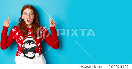 Winter holidays and celebration concept. Amazed redhead girl showing advertisement, pointing at christmas logo, standing excited over blue background 100623102