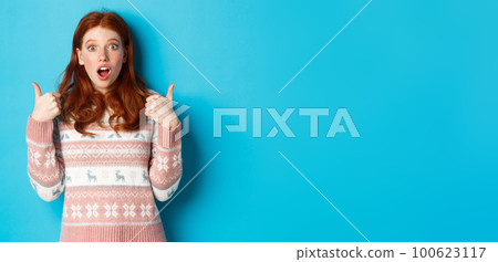 Impressed redhead girl in sweater showing thumbs-up, open mouth fascinated, approve and like product, praising something, standing over blue background 100623117