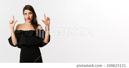 Fashion and beauty. Thoughtful attractive woman in black dress staring and pointing up, thinking with tempted look, white background Fashion and beauty. Thoughtful attractive woman in black dress staring and pointing up, thinking with tempted look, white background 100623131